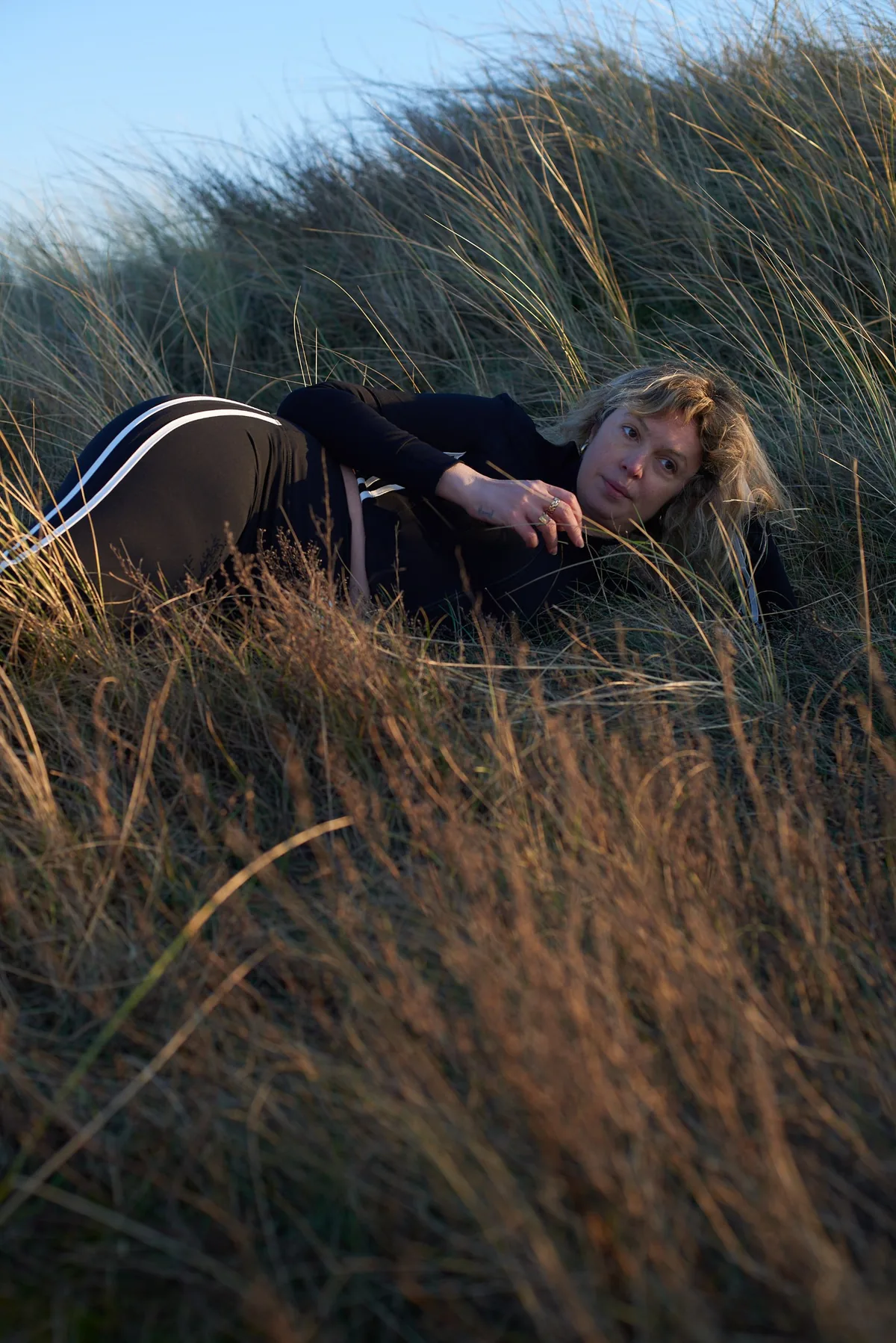 A Moontian lying in tall dune grass at sunset in the black GRAVITY LINE tracksuit, white stripes along the hip and leg