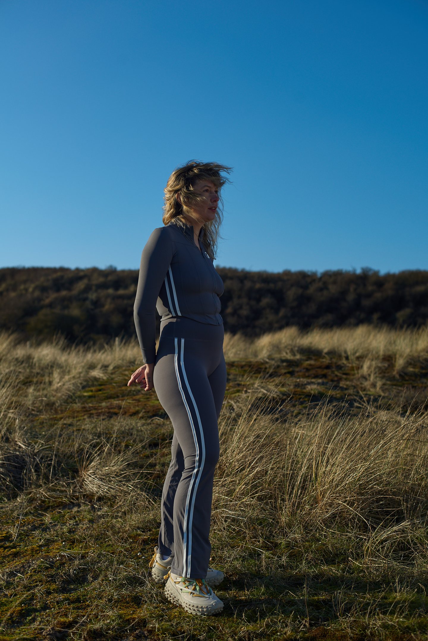A Moontian lying back in the dune grass in the grey two-stripe tracksuit, one arm across her brow
